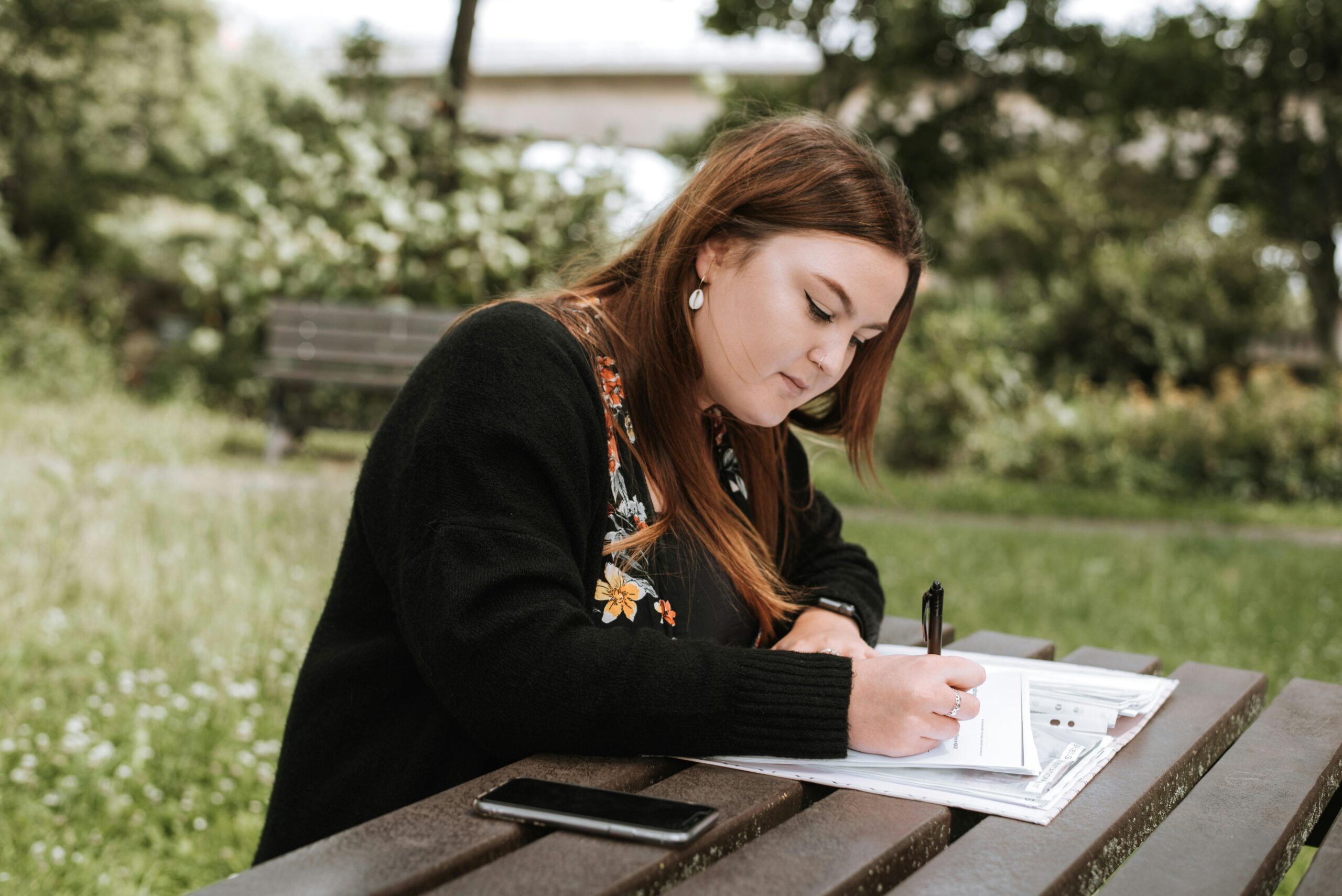 Focused young woman writing in a notebook while studying outdoors in a summer park.